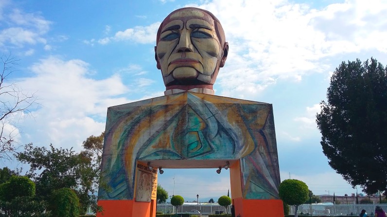  Gran escultura de la cabeza de Benito Juaréz, esta se encuentra encima de un arcos que sirve como entrada
                    