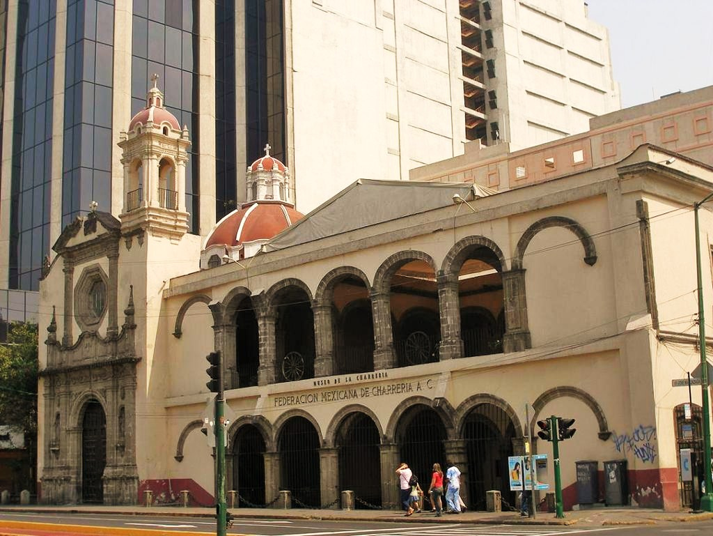 Fachada del museo de la charrería, Del lado izquierdo de la imagen se encuentra una iglesia y del lado derecho en adelante se encuentra el Museo con arcos de color gris formados por ladrillos y siendo la fachada color beige. 
                